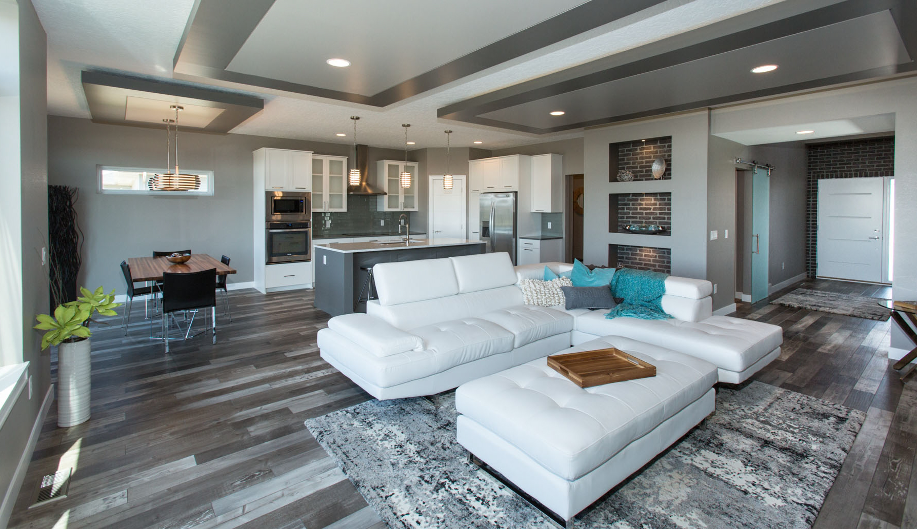 UPH open concept living, dining and kitchen area; white cabinets, grey tile backsplash, stainless steel appliances, grey island, pendant lighting over dining area and island, built-ins nad entry feature brick details; 8th St W